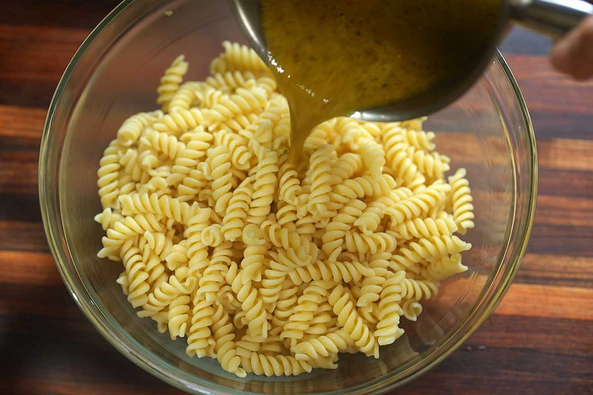 Warm dressing being poured over pasta and tossed to absorb