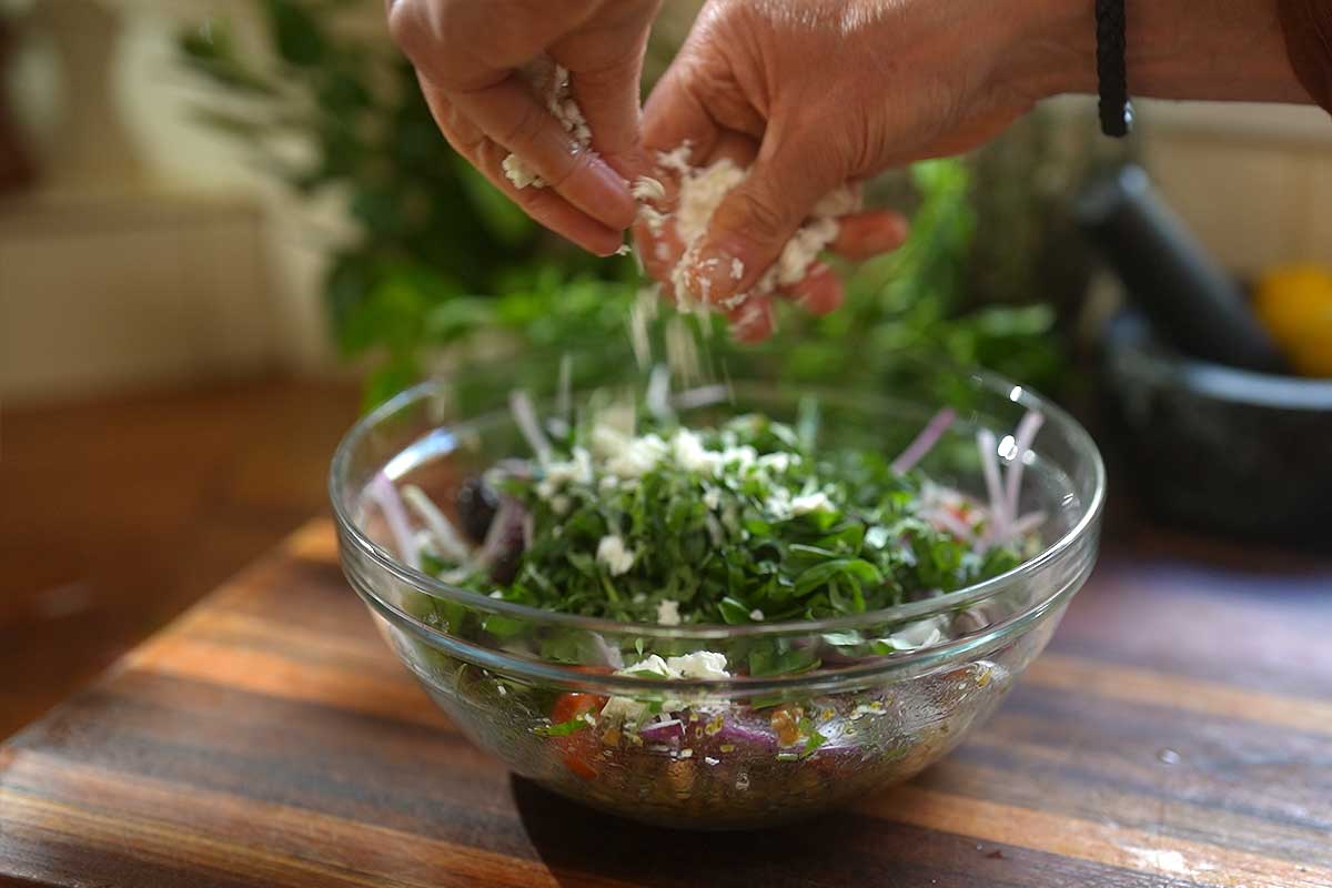 Salad being assembled with cherry tomatoes cucumber olives and crumbled feta