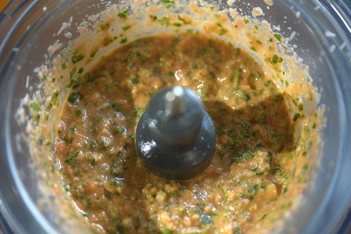 Close up of the coarse, textured pesto in the food processor bowl, showing visible pieces of almond and tomato