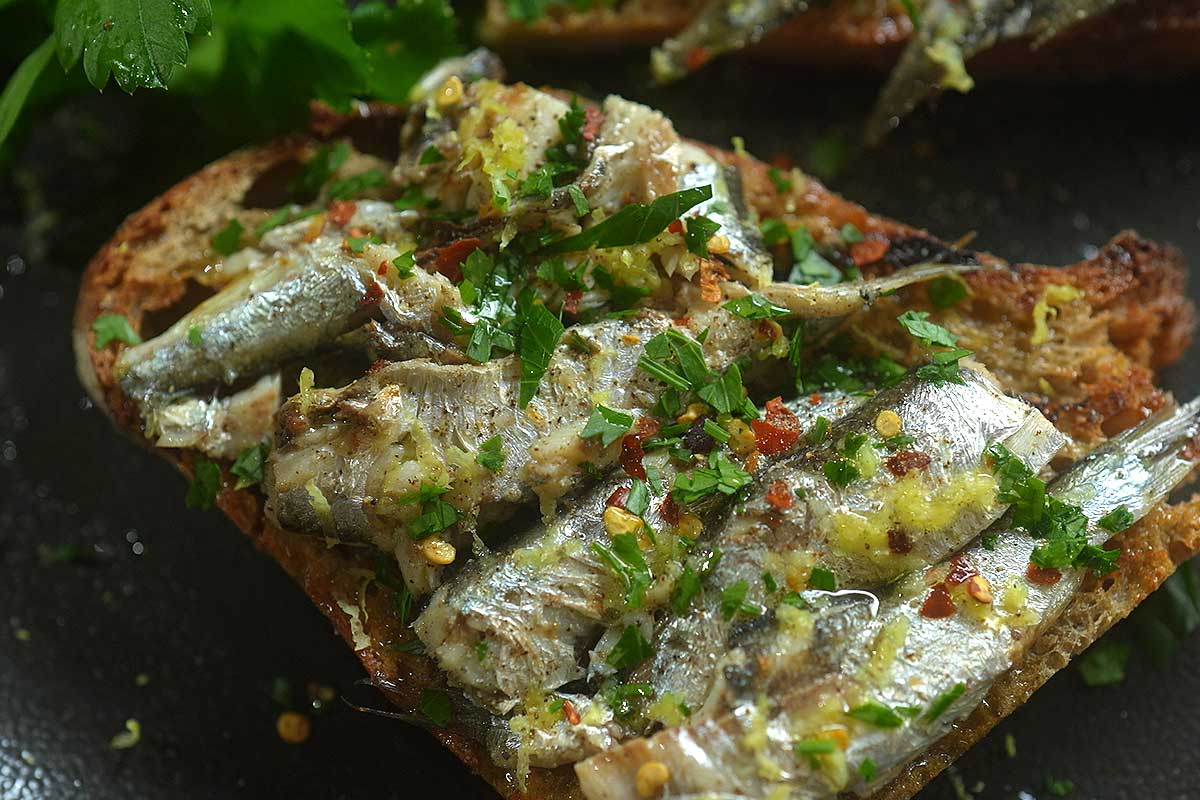 Overhead flat lay of two finished sardine toasts with lemon wedges and a small dish of chilli flakes