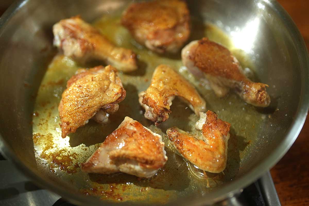 Chicken pieces browning skin side down in a large frying pan