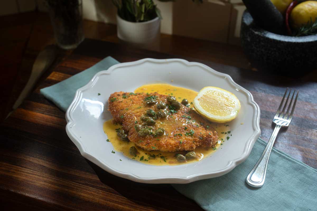 Overhead shot of the finished schnitzel piccata on a plate, sauce pooled underneath, lemon wedge on the side