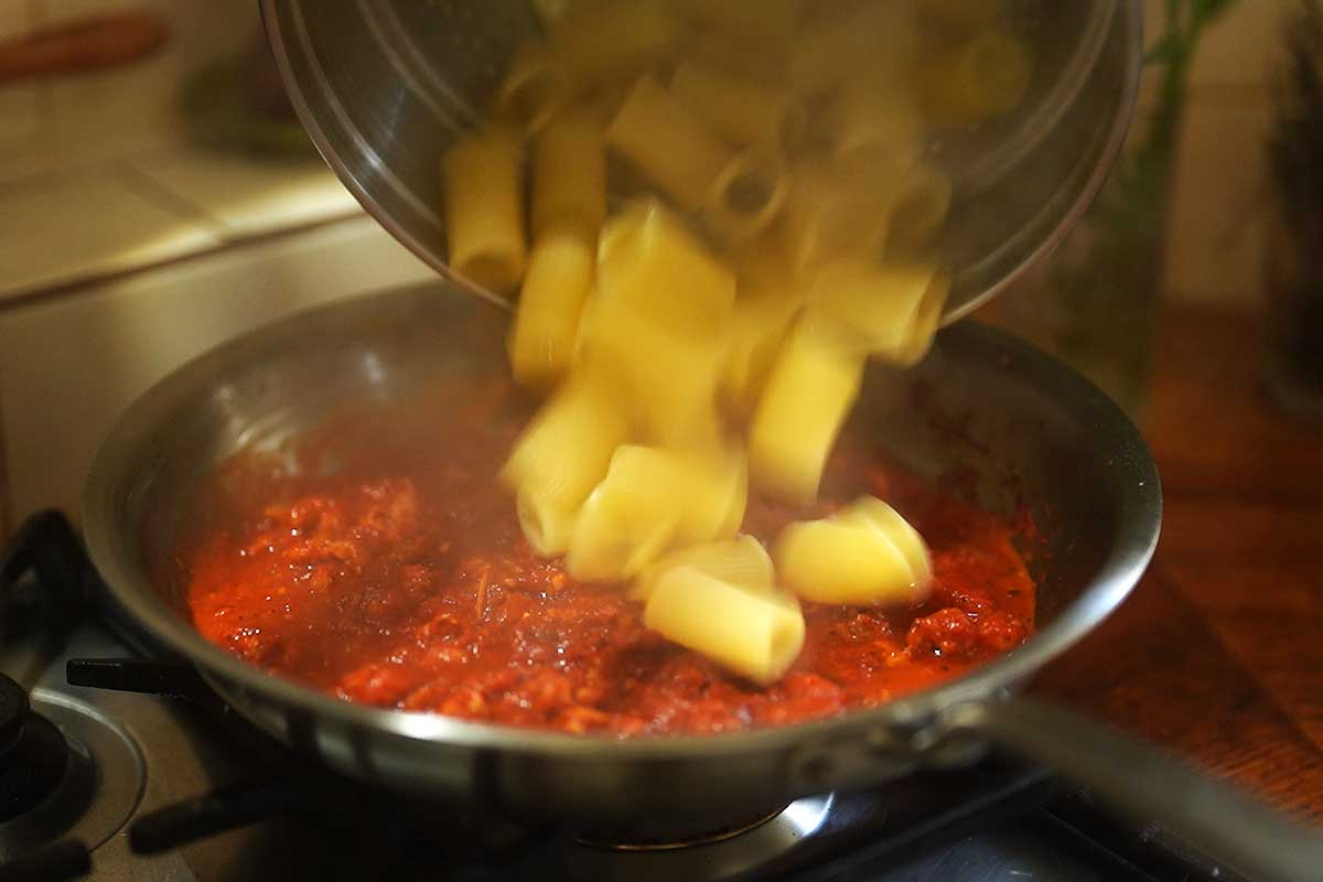 Drained rigatoni being tossed through the sauce with a splash of pasta water, turning glossy
