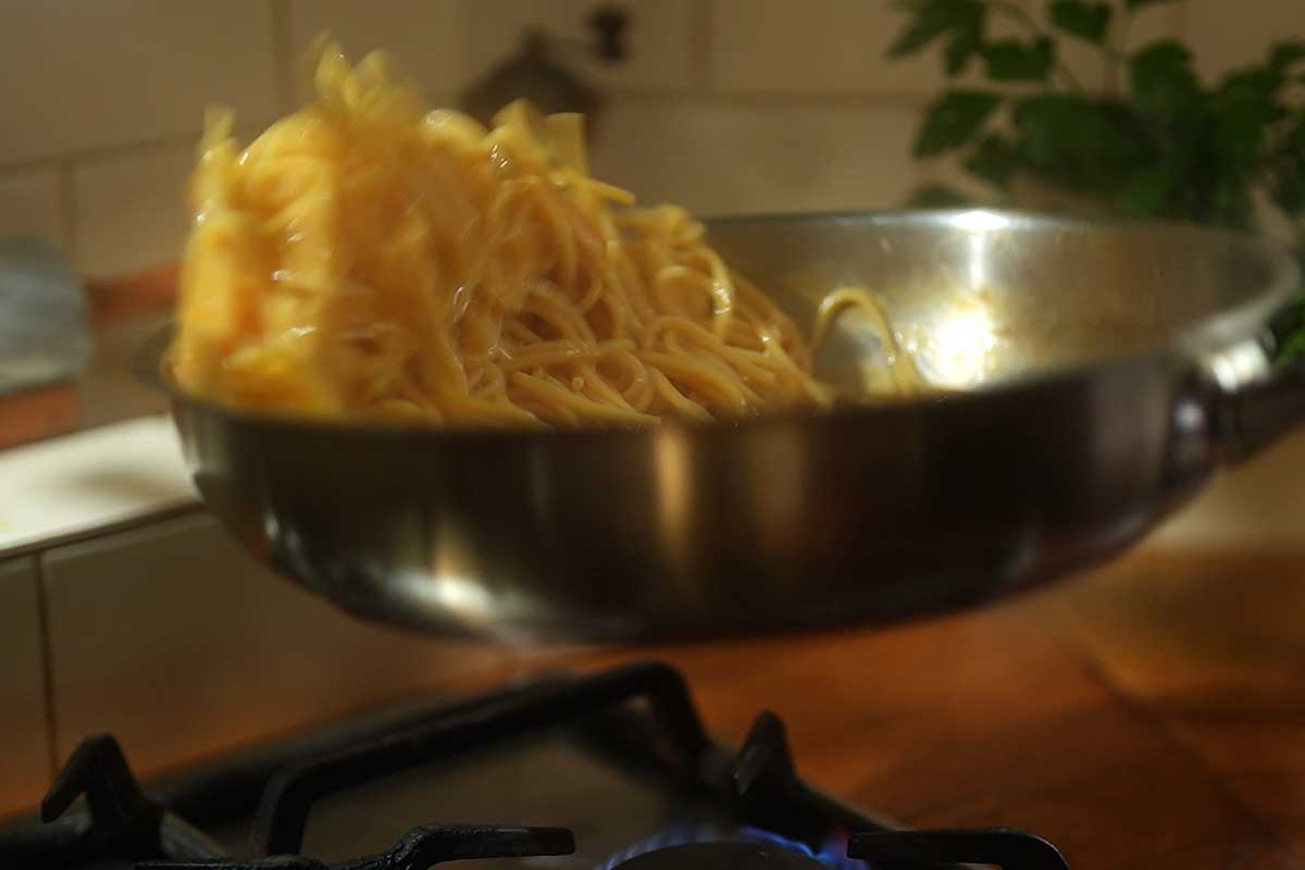Olive oil being drizzled into pasta while tossing vigorously to emulsify